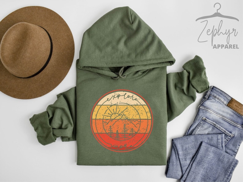 May include: A green hooded sweatshirt with a retro-style graphic of a mountain range, sun, and trees. The graphic is surrounded by a circle with the text "Explore More" in a vintage font.