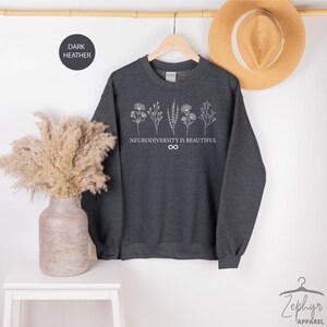 Op de afbeelding: Donkergrijze sweatshirt met een witte afbeelding van gestileerde bloemen en de tekst "Neurodiversity is beautiful" met een oneindigheidssymbool.