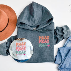May include: A dark grey hooded sweatshirt with the words "Pray Pray Pray" printed on the front in a colourful retro font. The back of the sweatshirt has the same text with the addition of "Do it, Over it, Through it" in smaller font. The sweatshirt is displayed on a white background with a brown hat and blue jeans.