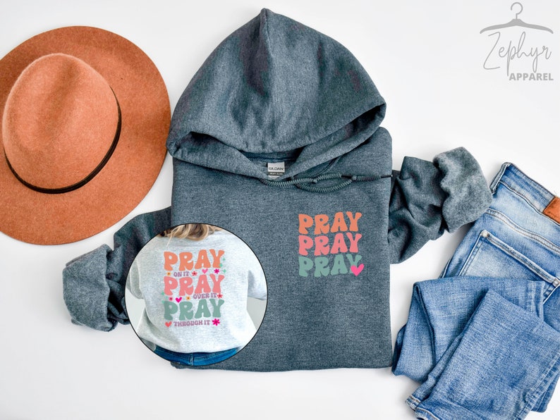 May include: A dark grey hooded sweatshirt with the words "Pray Pray Pray" printed on the front in a colourful retro font. The back of the sweatshirt has the same text with the addition of "Do it, Over it, Through it" in smaller font. The sweatshirt is displayed on a white background with a brown hat and blue jeans.
