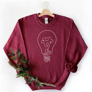 May include: A maroon sweatshirt with a white outline of a lightbulb with a puzzle piece inside. The lightbulb has a heart at the bottom of the filament.