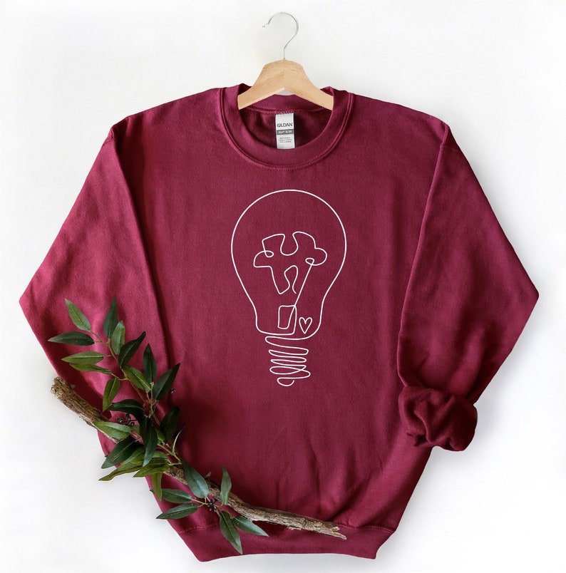 May include: A maroon sweatshirt with a white outline of a lightbulb with a puzzle piece inside. The lightbulb has a heart at the bottom of the filament.