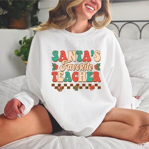 K&ouml;nnte beinhalten: Wei&szlig;er Pullover mit dem Text "SANTA'S FAVORITE TEACHER" in bunten Retro-Lettern mit einem karierten Muster und weihnachtlichen Akzenten.