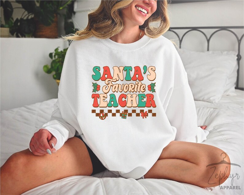 K&ouml;nnte beinhalten: Wei&szlig;er Pullover mit dem Text "SANTA'S FAVORITE TEACHER" in bunten Retro-Lettern mit einem karierten Muster und weihnachtlichen Akzenten.
