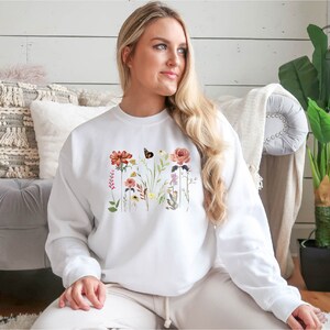May include: White sweatshirt with a floral print featuring a butterfly, red, pink, and yellow flowers, and green leaves.