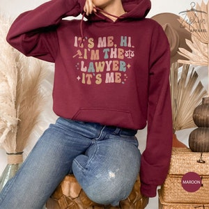 Peut inclure: Un sweat &agrave; capuche bordeaux avec le texte "It's me, hi. I'm the lawyer. It's me." en lettres color&eacute;es de style r&eacute;tro. Le sweat &agrave; capuche a un cordon de serrage et une poche kangourou.