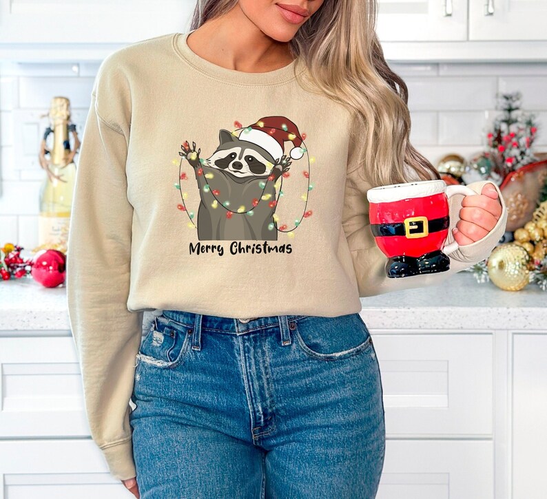 May include: A beige sweatshirt with a graphic of a raccoon wearing a Santa hat and holding Christmas lights. The text "Merry Christmas" is printed below the raccoon.