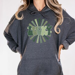 Peut inclure: Un sweat &agrave; capuche gris fonc&eacute; avec un motif vert et blanc du mot "LUCKY" dans une police r&eacute;tro avec un design en forme de soleil.