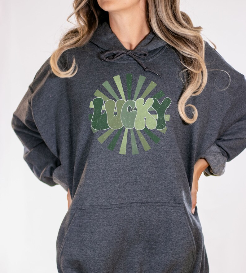 Peut inclure: Un sweat &agrave; capuche gris fonc&eacute; avec un motif vert et blanc du mot "LUCKY" dans une police r&eacute;tro avec un design en forme de soleil.