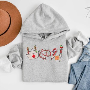 Puede incluir: Una sudadera con capucha gris con un dise&ntilde;o navide&ntilde;o que incluye un gorro de enfermera, un estetoscopio, vendas y una jeringa. El dise&ntilde;o es en rojo, blanco y verde.