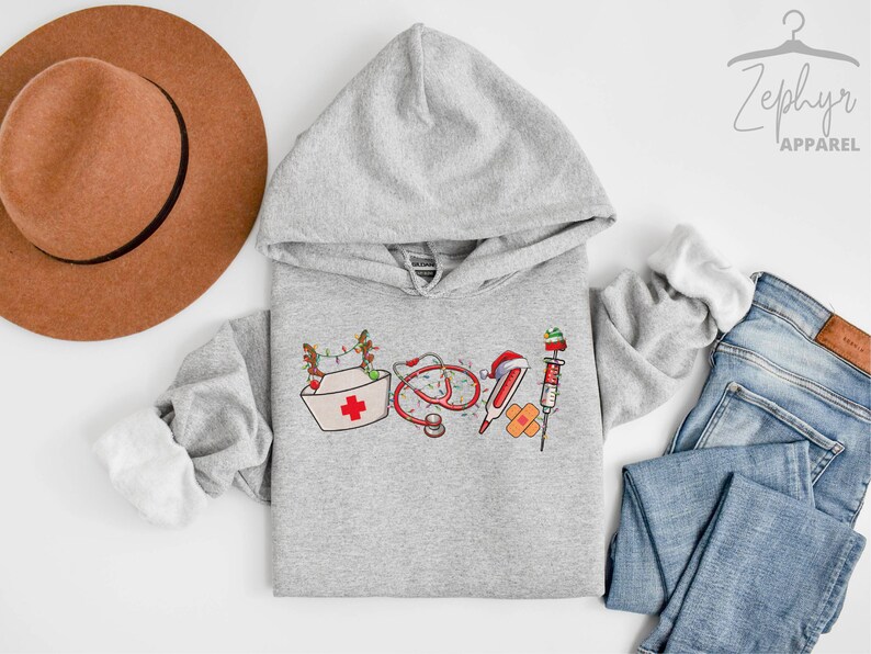 Puede incluir: Una sudadera con capucha gris con un dise&ntilde;o navide&ntilde;o que incluye un gorro de enfermera, un estetoscopio, vendas y una jeringa. El dise&ntilde;o es en rojo, blanco y verde.