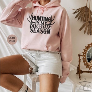 K&ouml;nnte beinhalten: Hellrosa Hoodie mit einem schwarzen Aufdruck, der "Hunting is my favorite season" mit Geweih und einem Zielsymbol zeigt.