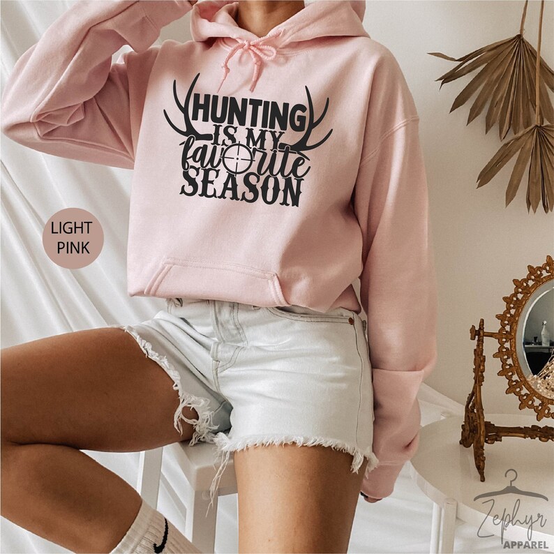 K&ouml;nnte beinhalten: Hellrosa Hoodie mit einem schwarzen Aufdruck, der "Hunting is my favorite season" mit Geweih und einem Zielsymbol zeigt.