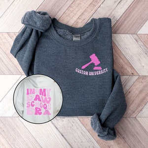 May include: A dark gray sweatshirt with a pink gavel and the text "Custom University" on the front. A white sweatshirt with the text "In My Law School Era" in pink letters is also shown.