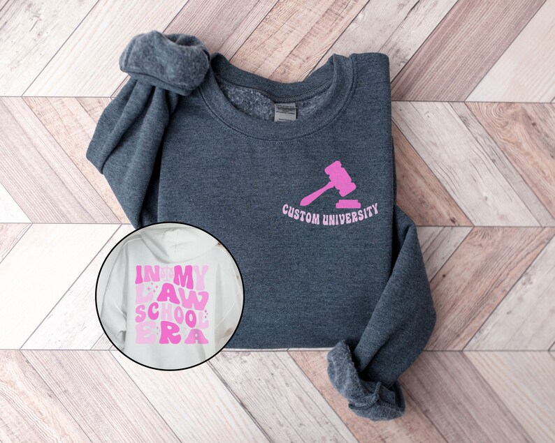 May include: A dark gray sweatshirt with a pink gavel and the text "Custom University" on the front. A white sweatshirt with the text "In My Law School Era" in pink letters is also shown.