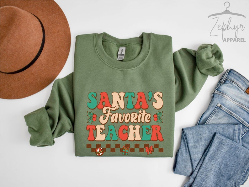 K&ouml;nnte beinhalten: Ein gr&uuml;ner Pullover mit dem Text "SANTA'S FAVORITE TEACHER" in einer Retro-Schriftart mit roten, orangen und t&uuml;rkisfarbenen Farben. Der Text ist von Stechpalmenbl&auml;ttern und roten Schleifen umgeben.