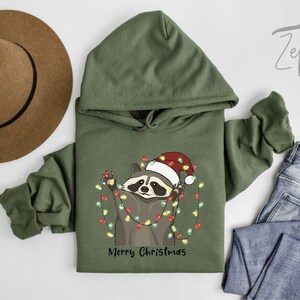 May include: A green hooded sweatshirt with a raccoon wearing a Santa hat and holding Christmas lights. The text "Merry Christmas" is printed below the raccoon.
