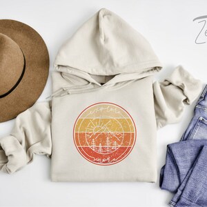 May include: A beige hoodie with a vintage-style graphic of a sun, mountains, and trees in a circle. The graphic is in shades of orange, yellow, and red. The text "explore" and "more" is written in a circle around the graphic.