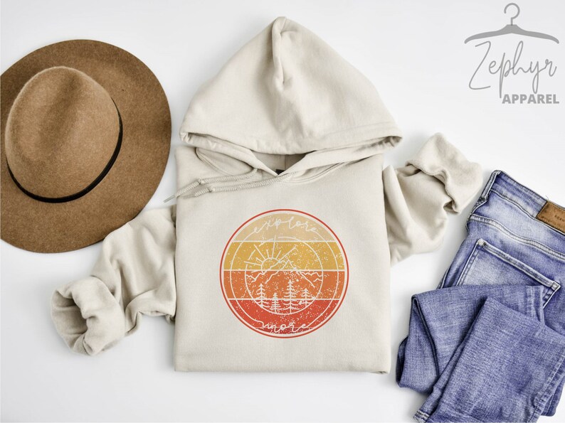 May include: A beige hoodie with a vintage-style graphic of a sun, mountains, and trees in a circle. The graphic is in shades of orange, yellow, and red. The text "explore" and "more" is written in a circle around the graphic.