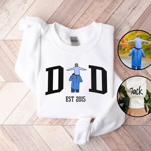 May include: White sweatshirt with a black graphic of a father carrying a child on his shoulders. The graphic says "DAD EST 2015".