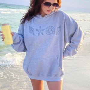 May include: A light gray hoodie with a front pocket and a seashell design. The design includes a seashell, starfish, and other sea creatures. A yellow can cooler is held in the hand. The person is standing on a beach.