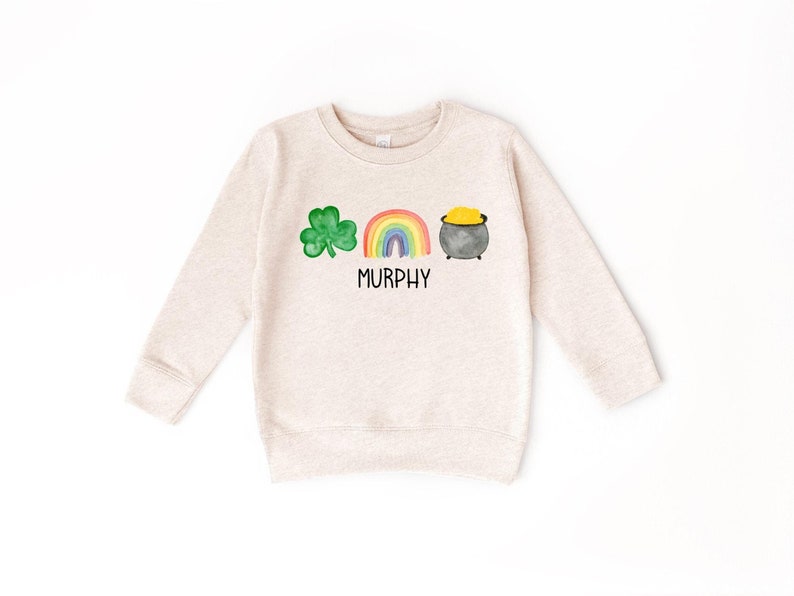 May include: A beige long-sleeved sweatshirt with a green shamrock, rainbow, and pot of gold graphic. The text "MURPHY" is printed below the graphic.