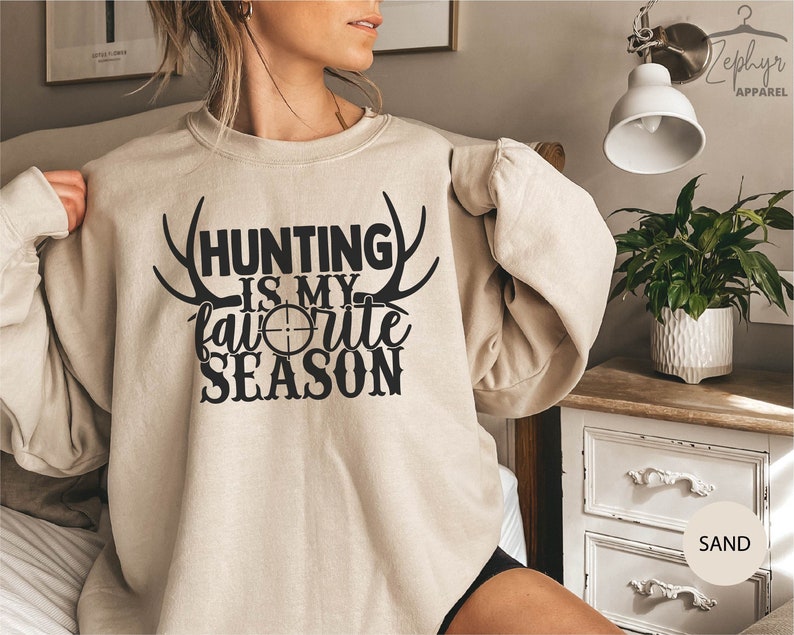 K&ouml;nnte beinhalten: Ein beiger Pullover mit dem Text "Hunting is my favorite season" in schwarzer Schrift mit einem Zielsymbol und Geweih.