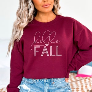Op de afbeelding: Bordeauxkleurige trui met een witte afbeelding die "hello fall" zegt met een klein hartje boven het woord "fall".