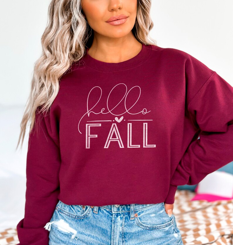 Op de afbeelding: Bordeauxkleurige trui met een witte afbeelding die "hello fall" zegt met een klein hartje boven het woord "fall".