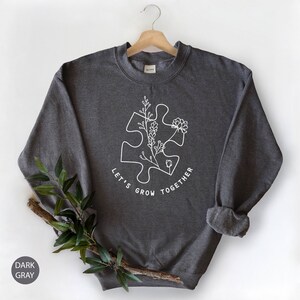 May include: Dark gray sweatshirt with a white outline of a puzzle piece with flowers growing from it. The text "Let's Grow Together" is written around the puzzle piece.