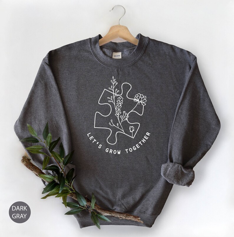 May include: Dark gray sweatshirt with a white outline of a puzzle piece with flowers growing from it. The text "Let's Grow Together" is written around the puzzle piece.
