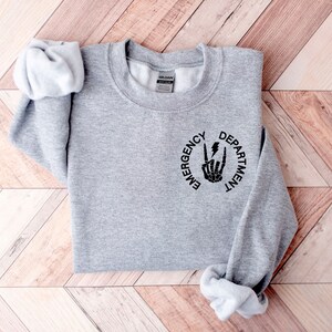 May include: Gray sweatshirt with a black graphic of a skeleton hand making a rock and roll sign with a lightning bolt. The graphic is inside a circle with the words "Emergency Department" around it.