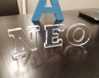 May include: Clear acrylic letters spelling out the word "NEO". The letters are standing on a dark wood surface.