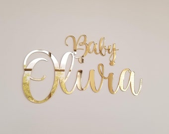 Może przedstawiać: Złoty akrylowy znak z imieniem dziecka z lustrzanym wykończeniem z tekstem "Baby Olivia" w ozdobnym, pochylonym piśmie.