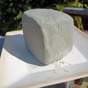 May include: A block of gray clay, roughly rectangular with rounded edges, sits on a white ceramic plate. The clay has a textured surface and appears to be freshly molded. The plate has a slightly raised rim and a subtle, warm color.