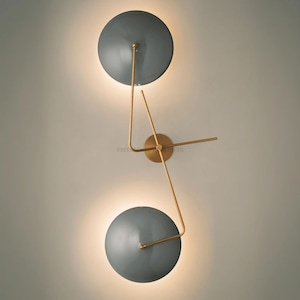 May include: A modern wall sconce with two round, gray shades and gold metal arms. The shades are angled and the arms are connected to a central base.