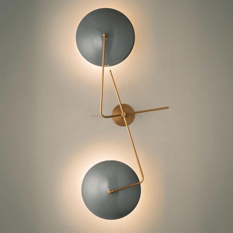 May include: A modern wall sconce with two round, gray shades and gold metal arms. The shades are angled and the arms are connected to a central base.