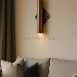 May include: A modern wall sconce with a cylindrical bronze-colored light fixture, mounted on a black diamond-shaped base. The sconce is illuminated, casting a warm glow. The text "VINTAGENAUTICALGIFTS" is visible. The sconce is above a beige sofa with pillows.