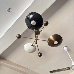 Könnte beinhalten: Eine moderne Deckenleuchte mit fünf Armen und vier Schirmen. Die Schirme sind schwarz, weiß und goldfarben. Die Leuchte ist mit einer goldenen Stange an der Decke befestigt.
