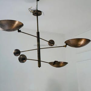 Könnte beinhalten: Ein Messing-Kronleuchter mit fünf Lichtern. Der Kronleuchter hat ein einzigartiges Design mit mehreren Armen, die sich von einer zentralen Stange erstrecken. Jeder Arm hat eine runde Leuchte und einen geschwungenen, schalenförmigen Schirm.
