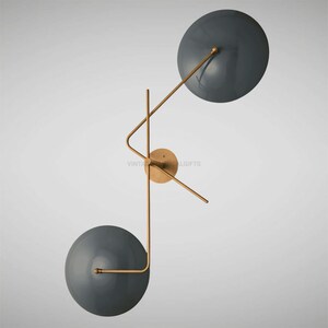 May include: A modern wall sconce with two round, dark gray shades. The shades are attached to a gold metal frame with a geometric design.