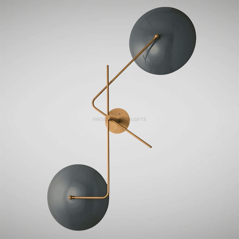 May include: A modern wall sconce with two round, dark gray shades. The shades are attached to a gold metal frame with a geometric design.