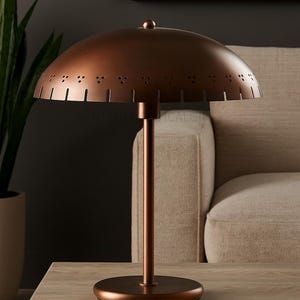 May include: A copper-colored table lamp with a domed shade featuring decorative cutouts. The lamp has a slender stem and a circular base. It is placed on a wooden table, with a neutral-toned sofa in the background.