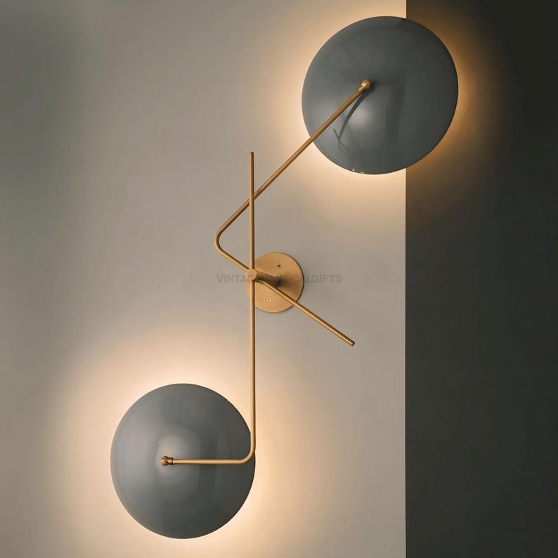 May include: A modern wall sconce with two round, gray shades and a gold metal frame. The shades are angled in different directions, creating a unique and stylish design.