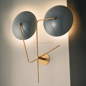 May include: A modern wall sconce with two gray, round shades and a gold metal frame. The shades are angled outward and the frame is attached to the wall with a circular base.