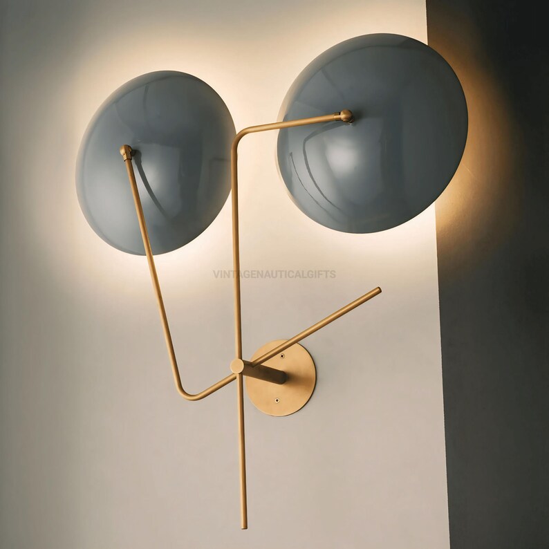 May include: A modern wall sconce with two gray, round shades and a gold metal frame. The shades are angled outward and the frame is attached to the wall with a circular base.
