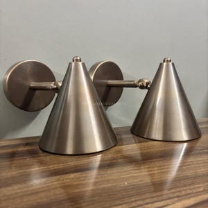 Op de afbeelding: Een paar wandlampen in de vorm van een kegel van geborsteld nikkel met een modern ontwerp. De lampen zijn aan een muur gemonteerd en hebben een eenvoudige, elegante uitstraling.