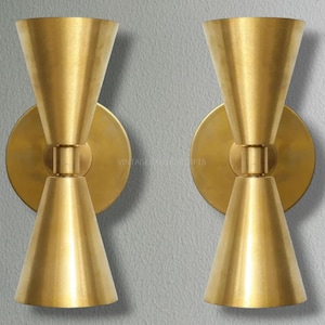 Op de afbeelding: Een paar goudkleurige wandlampen met een uniek zandloperontwerp. Elke lamp heeft twee verstelbare kegelvormige lampenkappen.