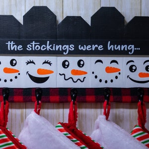 May include: A wooden stocking holder with a black and red plaid base and five snowman faces painted on the front. The top of the holder has a black sign that reads "the stockings were hung..."