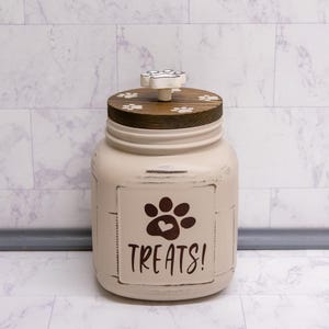 May include: A beige ceramic jar with a distressed finish, featuring a dark brown paw print with a heart center and the text TREATS! in black. The jar has a dark wood lid with white paw prints and a white paw-shaped handle on top.
